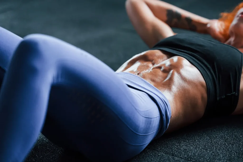 Abdominaux ciselés : séance intense pour un ventre sculpté Femme rousse musclée faisant des abdominaux, ventre très ciselé et luisant de sueur, vêtue d'un legging bleu et d'un haut noir.