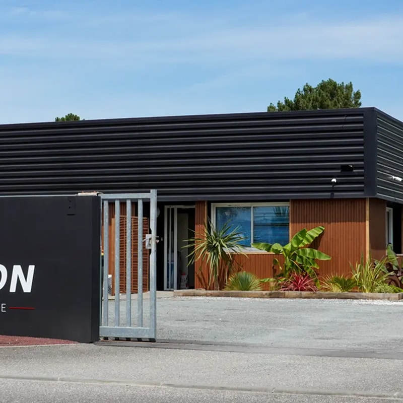 Entrée d'un bâtiment moderne avec façade marron et toit noir ondulé, signalétique 'ON' visible, et portail métallique ouvert sur une cour en gravier.