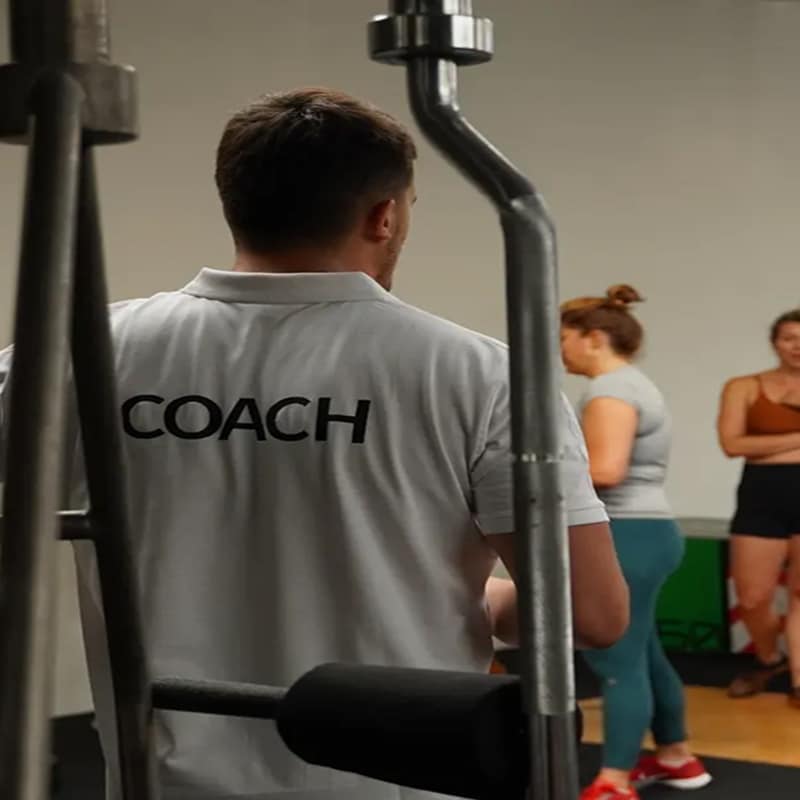 Vue de dos d'un coach portant un polo blanc avec l'inscription "COACH" en noir, encadré par un appareil de musculation, face à des femmes dans une salle de sport.