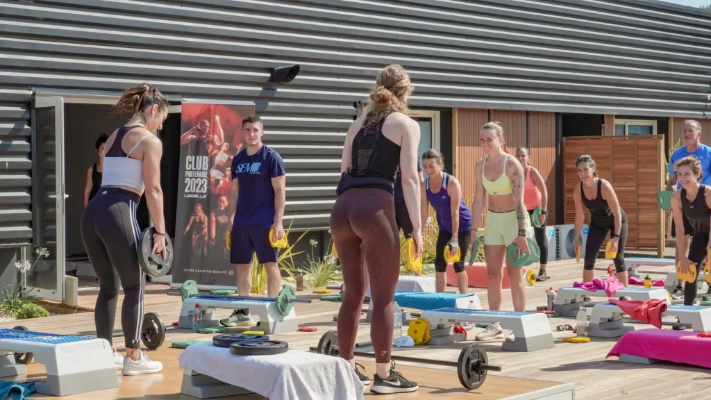 Cours de fitness Les Mills en extérieur avec haltères Groupe de sportifs effectuant un entraînement Les Mills avec haltères et step, un homme affiche le poster "CLUB PARTENAIRE 2023".