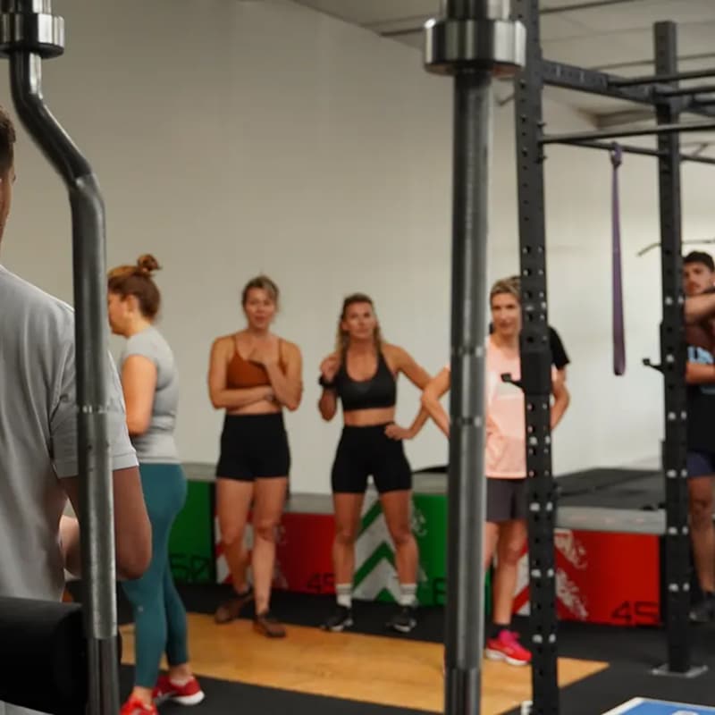 Un groupe de cinq femmes et un homme écoutent un homme en gros plan lors d'une séance d'entraînement en salle de sport avec des racks de musculation.