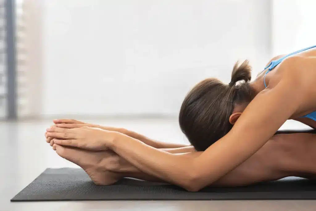 Étirement Jambes Assis: Flexion Avant sur Tapis Jeune femme brune en haut bleu effectue un étirement assis, se penchant vers l'avant pour saisir ses pieds sur un tapis de yoga noir.