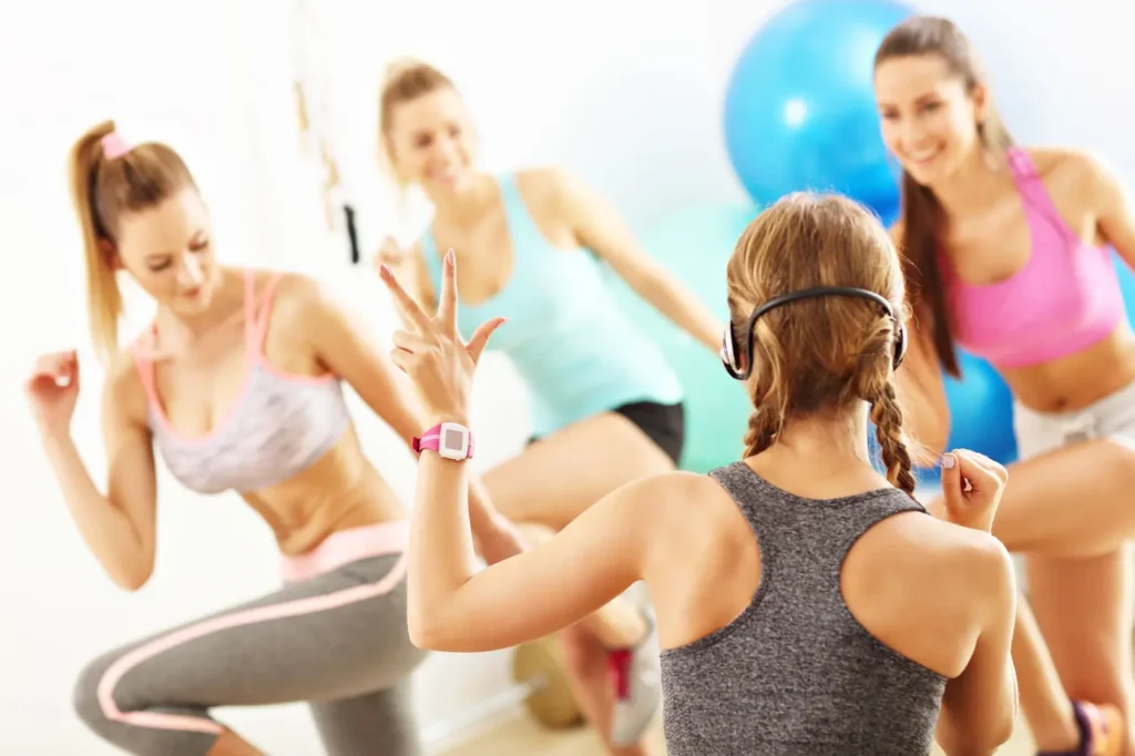 Cours de fitness cardio : femmes souriantes en pleine séance d'entraînement Quatre femmes pratiquent un exercice de fitness ou de danse cardio. L'une d'elles porte un tracker d'activité rose et fait le signe de la paix avec les doigts.