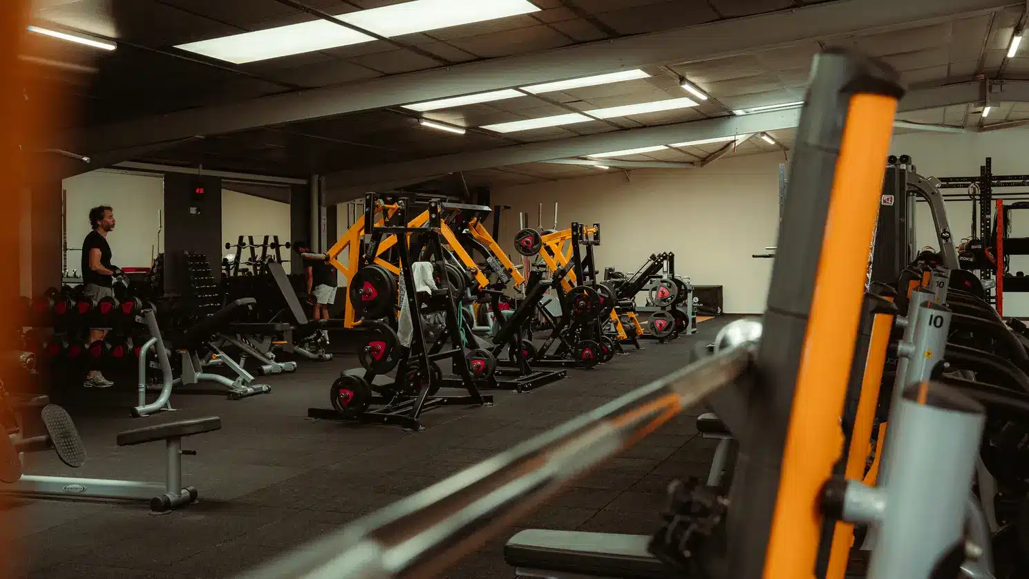 Machines de musculation orange et noir dans une salle de sport sombre. Un athlète se prépare près du rack d'haltères. Horloge : 12:55.