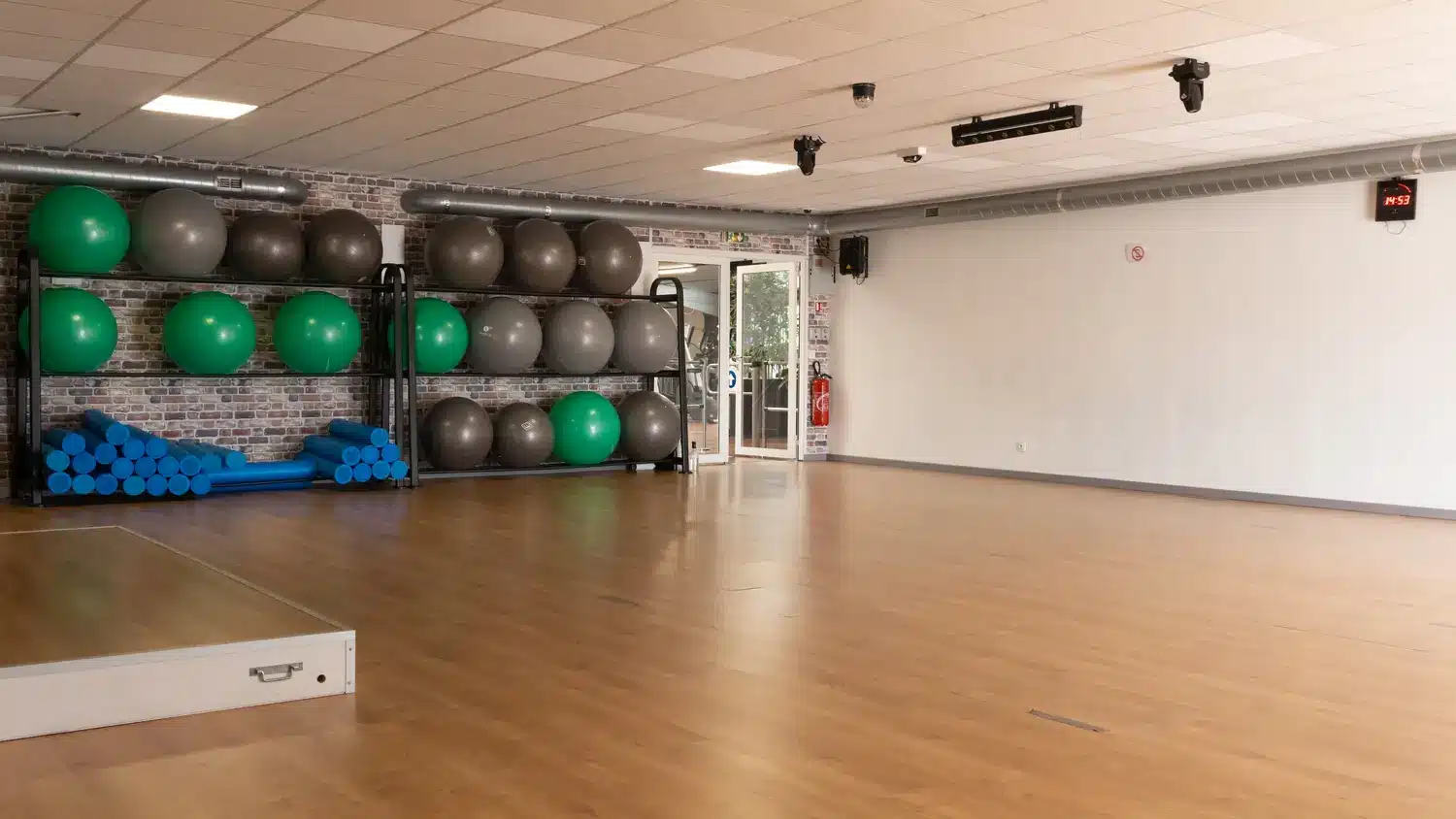 Salle de sport fonctionnelle et lumineuse, équipée de ballons de stabilité (verts et gris), rouleaux de mousse bleus et d'un plancher en bois.