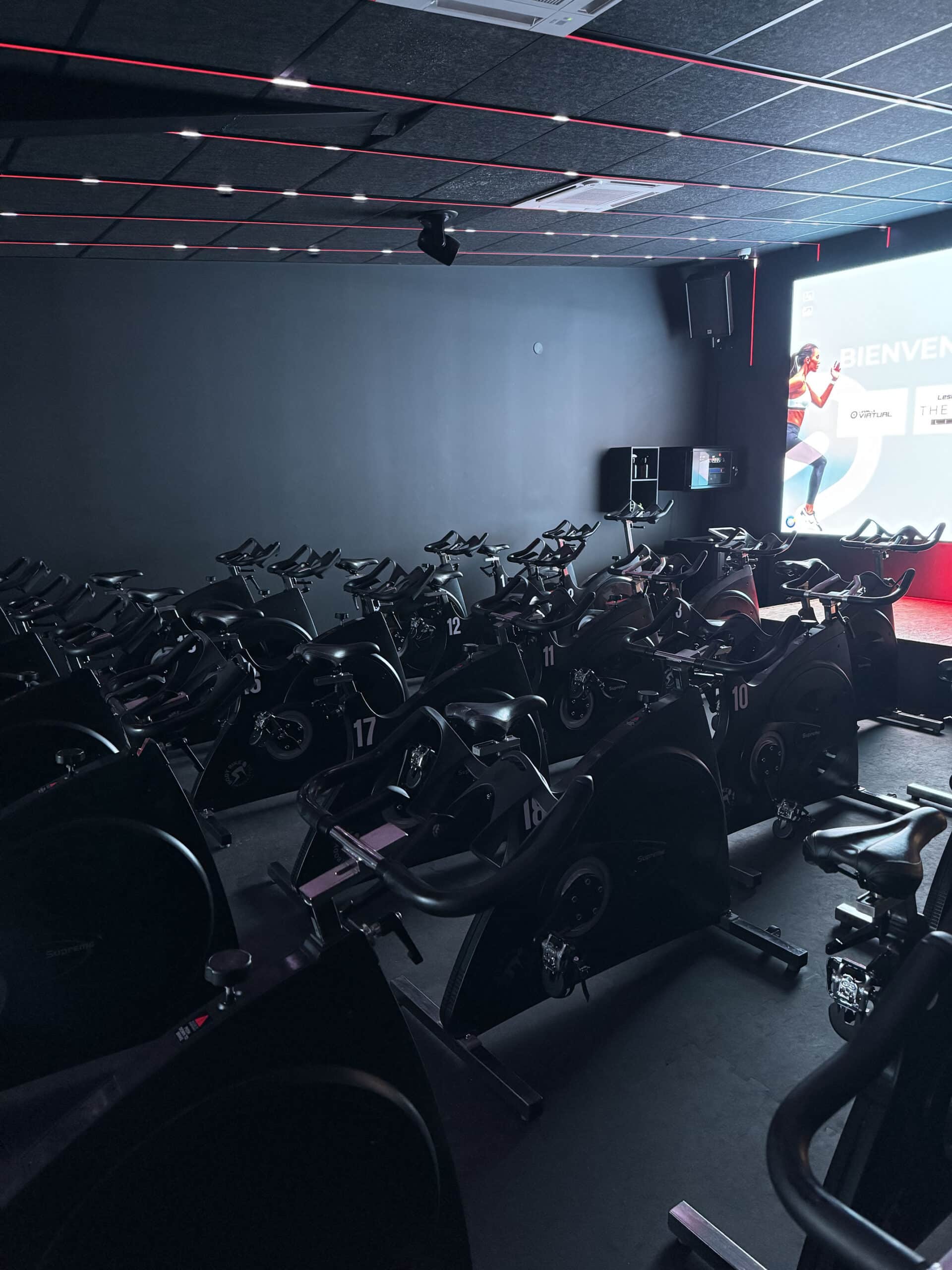 Salle de RPM sombre avec des rangées de vélos stationnaires numérotés. Lignes LED rouges au plafond. Écran affichant "BIENVENUE" et cours de cycling virtuel.